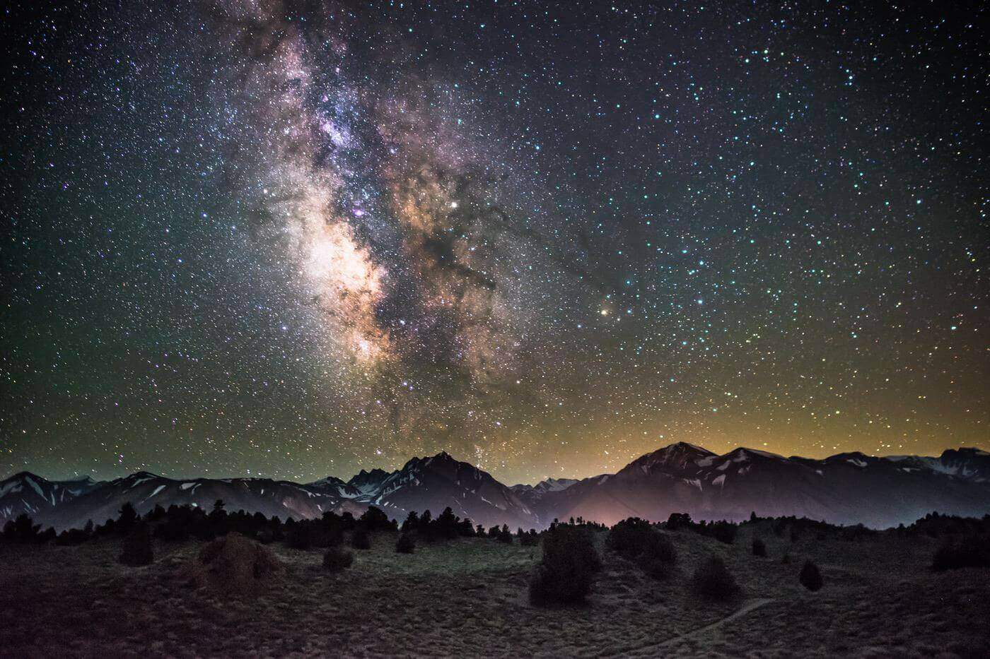 The milky way above a foggy landscape with mountains in the background and a forest in the foreground.